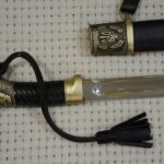 Cossack shashka grip, wrist strap and fullered blade base beside the trident-decorated scabbard mouth