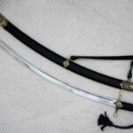 Curved SiCh-Hetman blade beside its black scabbard, showing the patterned scabbard tip and carrying strap